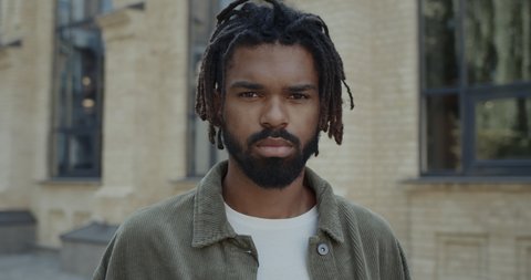 Portrait of mixed race guy with beard looking to camera. Close up view of young serious man with dreadlocks standing at city street. Concept of lifestyle, headshot.