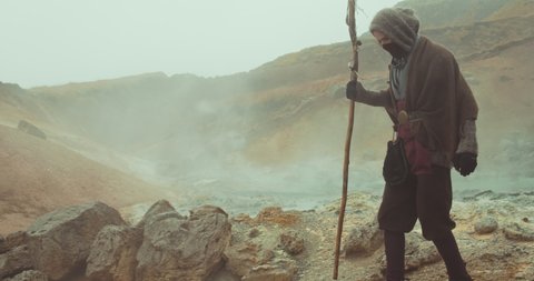 Handheld Slow Motion Wide Tracking Shot Of Hooded Man In Robes Carrying Staff Through Misty Desert Landscape And Stopping To Look Around