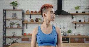 Young woman blogger with short colorful red and black haircut wearing blue top poses for camera in light contemporary kitchen