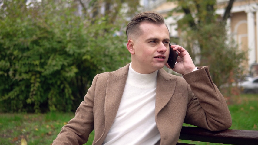 Elegant smiling man talking on the phone while sitting on a bench in the street