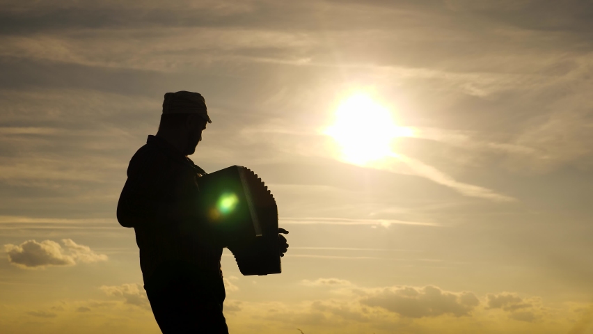 Man plays accordion in sun. Artist performs in open air, sings and plays musical instrument. Tradition and celebration in countryside. silhouette of cheerful man, he sings and plays musical instrument Royalty-Free Stock Footage #1064425423