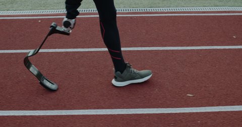 Side view of disabled male person legs with prosthetic running blade walking at sports field. Handicapped male sportsman getting ready for run. Concept of motivational sports footage