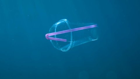 Plastic cocktail cup with a plastic straw slowly drifts underwater in blue water column in sunrays. Plastic garbage environmental pollution problem in seas and ocean. Adriatic Sea, Montenegro, Europe