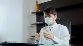 A young man in a medical mask sitting at the table disinfects his hands. Distance learning online.