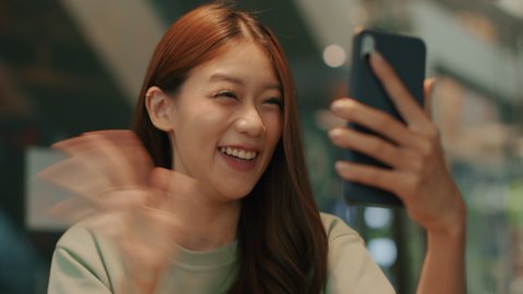 Young Beautiful Asian woman using a smartphone video calling, chatting online with her friend, family sitting in a cafe. Asian Girl waving hand and talking online communication on a mobile phone.
