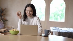 Portrait of young Asian woman in warm knitted clothes watching tv video movie series, online reality show on laptop computer, take a break while sitting on desk and eating popcorn in bed room at home.
