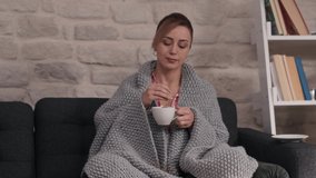 Sick woman sniffing herbal tea. Sick young woman trying to warm up with hot tea, wrapped in a blanket, resting on the sofa at home, coughing. Coronavirus. Health concept.