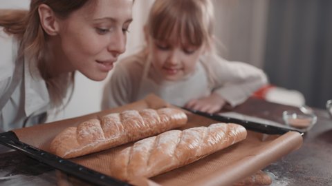 Medium close-up of joyful mom and daughter smelling two fresh loaves of bread lying on pan just baked in oven
