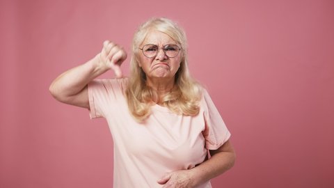 Disapproval concept. Grumpy senior woman looking at camera and gesturing thumb down gesture, pink studio background