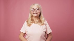 Love and charity. Positive adorable senior woman in glasses showing heart gesture near her chest, expressing kindness, pink studio background