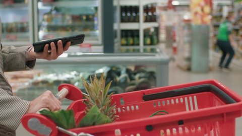 Midsection footage of unrecognizable woman using smartphone while carrying red shopping cart with fruit walking through aisles in hypermarket doing daily food shopping