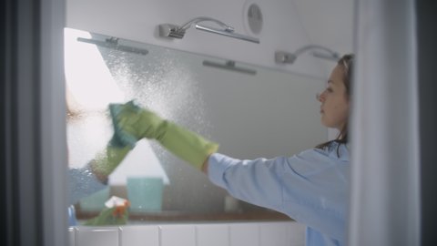 Young woman in bathroom wash the mirror with a spray for crystal clean it with a green rag