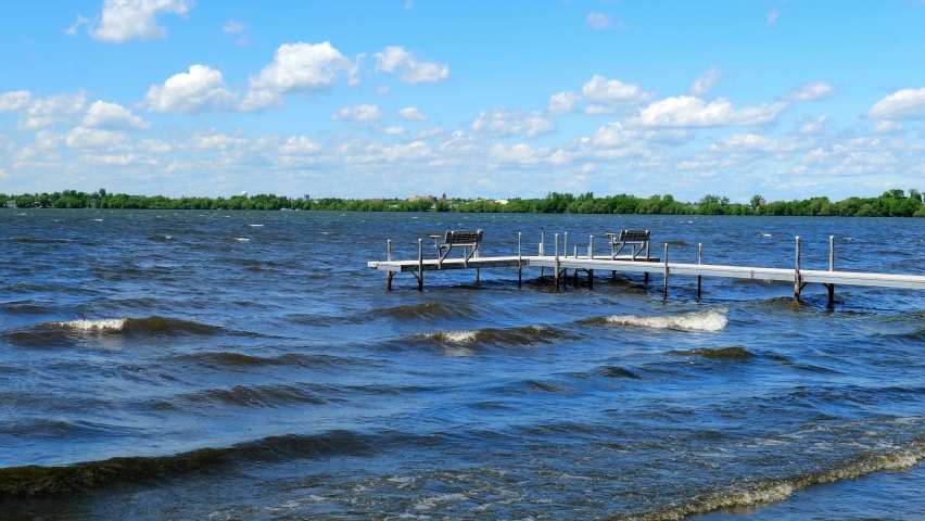 Boat dock with empty benches on Lake Irving, the first lake on the Mississippi River, with white waves rolling onto the beach in the city of Bemidji, Minnesota on a windy day with mostly blue sky.