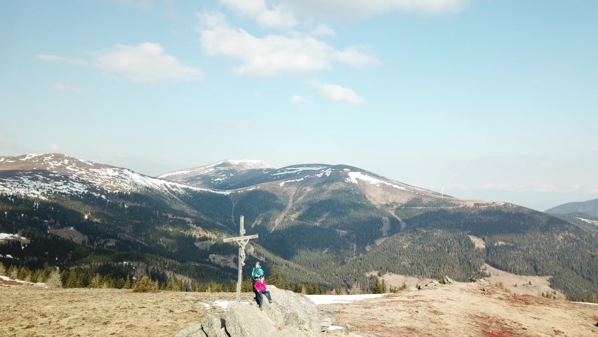 A drone capture of a couple with backpacks standing at the top of Seiner Kreuz, a peak in Austrian Alps. A wooden cross on the peak. Early spring in the mountains. Wanderlust. Achieving the goal