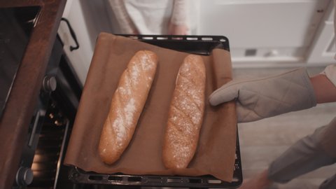 Top-view footage of unrecognizable little girl opening oven door and woman in oven mitt taking out baking sheet with two fresh loaves of bread