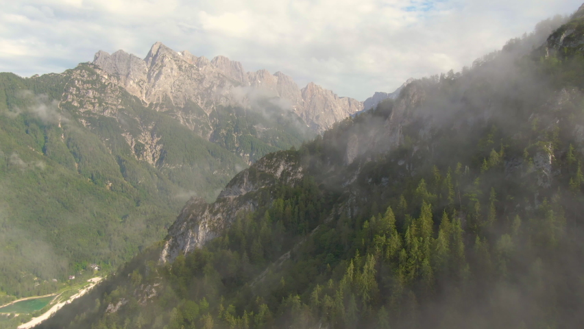 DRONE: Breathtaking drone view of the beautiful Julian Alps on a sunny summer day. Mist hovers over the coniferous woods covering the Julian Alps. Flying up a mountains in the picturesque Karawanks.