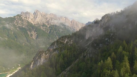 DRONE: Breathtaking drone view of the beautiful Julian Alps on a sunny summer day. Mist hovers over the coniferous woods covering the Julian Alps. Flying up a mountains in the picturesque Karawanks.