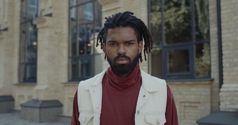 Portrait of serious guy with beard raising head and looking to camera. Crop view of young man with dreadlocks wearing turtleneck standing at city street. Zoom in. Concept of lifestyle.