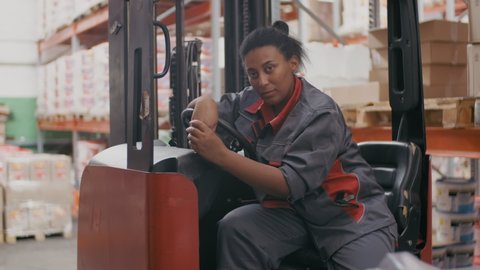 Medium slow-motion portrait of female african american warehouse worker in construction uniform looking at camera sitting in forklift working in big stockroom