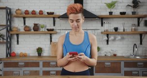 Cheerful lady blogger with stylish haircut in comfortable sportswear works surfing internet with phone in spacious kitchen