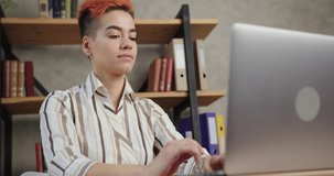 Attractive woman in stylish shirt has videocall on modern laptop sitting at table near bookshelves and beige wall aloneing home