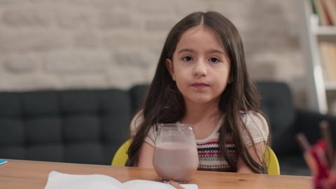Little girl taking a break from studying, laughing, looking at the camera. Drinking cocoa milk. Distance education of children in quarantine.Slow motion video.