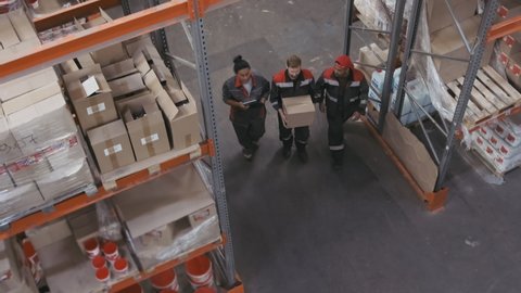 From-above shot of three multi-ethnic working people in coverall uniform clothes walking along pallet racks full of carton boxes and construction materials