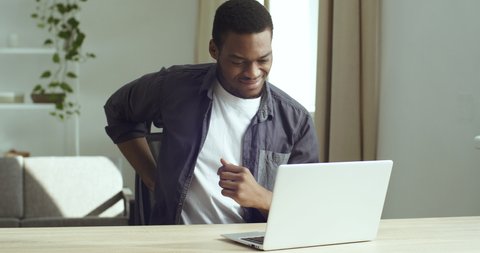 Afro american business man touching back sitting at desk feeling sudden backache pain, black tired sad guy suffering from incorrect bad posture work at laptop computer, rheumatism injury problems
