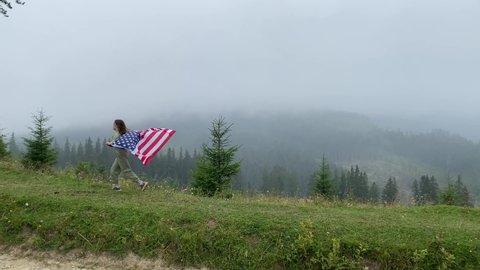 happy little girl is running and jumping carefree with open arms in cloudy mountains. Holding USA flag. selective focus.