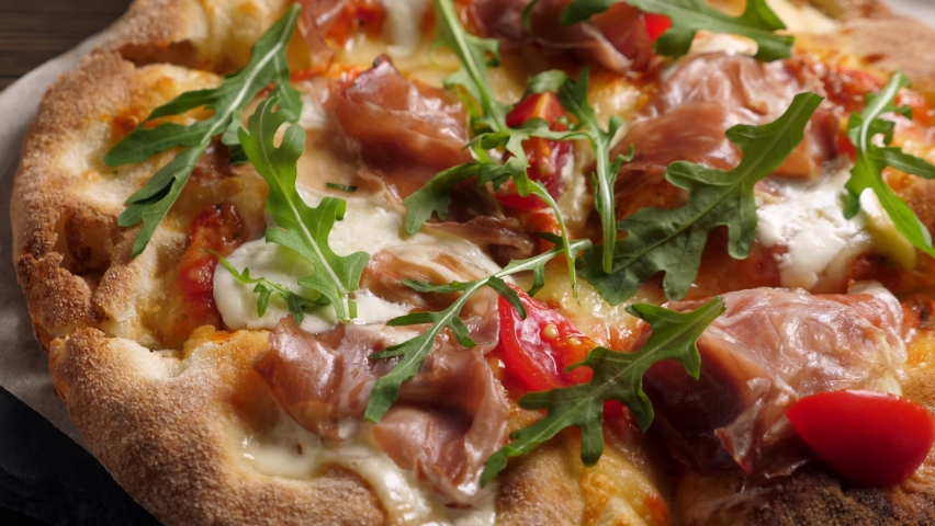 Prosciutto pizza with strachatella cheese, tomatoes and arugula leaves. Rotating on wooden table | Shutterstock HD Video #1064496424