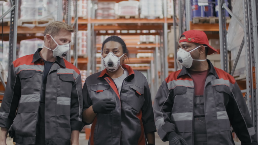 Medium front-view footage of group of multi-ethnic construction working people in uniform and protective masks walking towards camera along big warehouse with pallet racks, an Industrial Video by Pressmaster