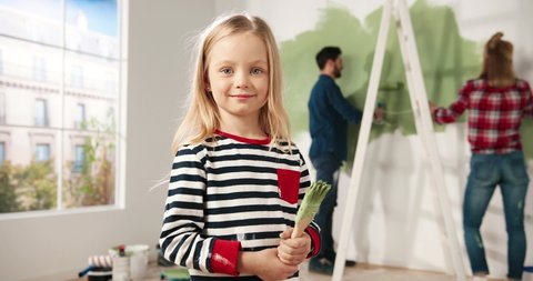 Close up portrait of adorable happy little cute girl smiling to camera standing in room with paint brush in hands. Mom and dad painting wall in green colour on background. Home repair. Interior design