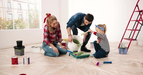 Caucasian joyful parents in new apartment with little cute girl speaking preparing paint for painting walls. Pretty preschool daughter child helps to her mom and dad to redesign house. Home repair