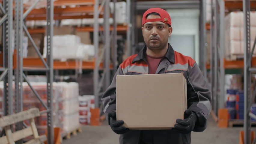 Medium footage of serious middle-eastern warehouse worker in blue coverall walking towards camera with big carton box along pallet racks | Shutterstock HD Video #1064497585