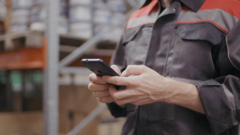 Midsection slow-motion shot of unrecognizable hands of workman in construction overall suit holding smartphone texting messages while walking at warehouse