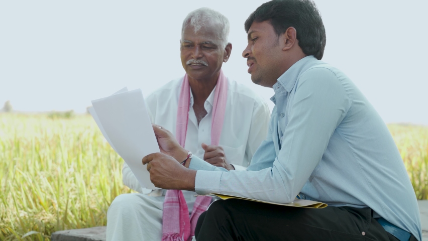Bank officer explaining about farm loan paper documents to Indian farmer near agriculture field - concept of Indian villager banking and lifestyle