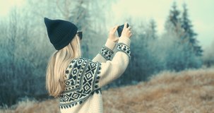 Woman traveler with her phone camera taking pictures beautiful winter snowy forest view. Fashion girl in wool sweater. Happy winter holidays. New Year's Eve. Christmas holiday. 4K video Slow motion.