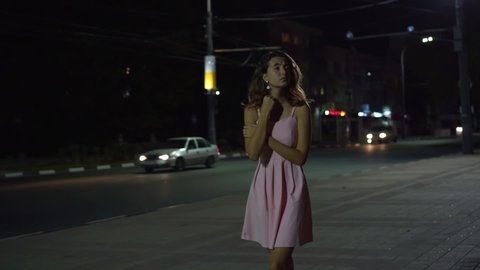 A lonely girl walks around the city at night, she froze and she is sad. A girl in a pink dress with curly hair is walking down the street at night in a state of hopelessness. Threw the guy.