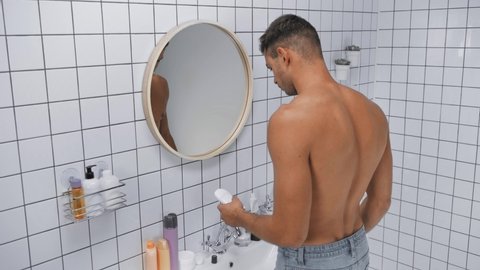 shirtless man applying deodorant while holding solid stick in bathroom