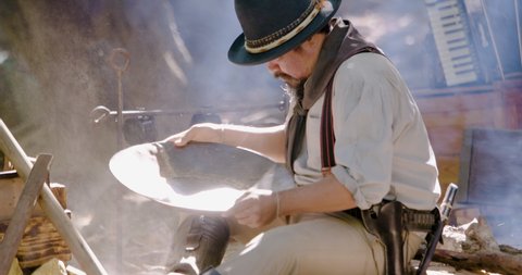 Gold digger wearing on cowboy hat and cowboy boots sitting using gold panning to refining the gold metal particles  while boiling the coffee flask above fire and some are play revolver gun