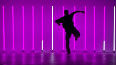 A young guy performs complex acrobatic figures in the studio against the background of multicolored neon lamps. Hip hop. Youth subculture. Contemporary choreography. Silhouette. Slow motion.