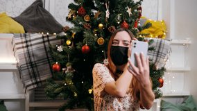 Cute girl under a decorated Christmas tree in a medical mask celebrates Christmas in quarantine via a video call from a smartphone with family and friends