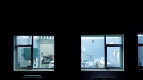 Operating rooms (operating theaters) in a hospital with a group of surgeons and nurses performing the surgery. Aerial view from outside through a window at night.