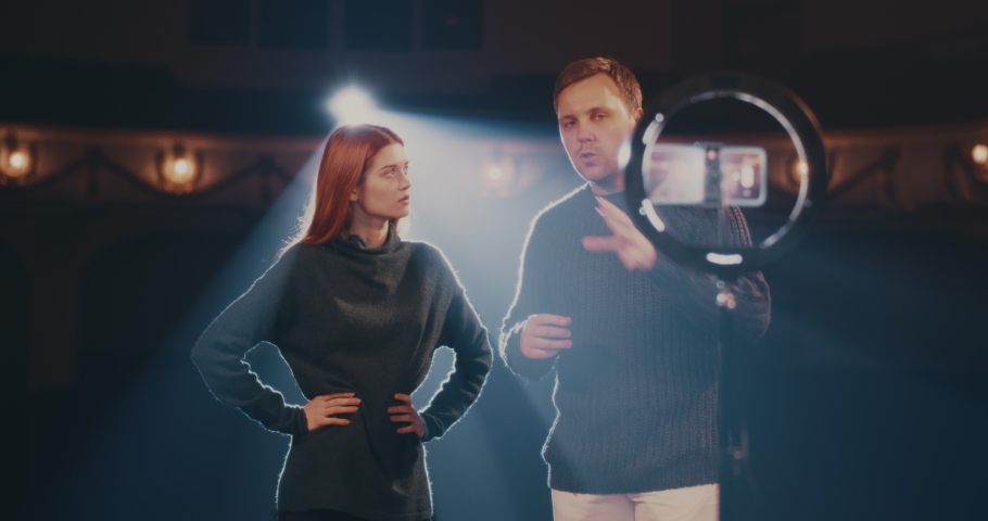 Adult man talking and young woman demonstrating stage smile while recording or streaming online tutorial video in spotlight on stage in theater