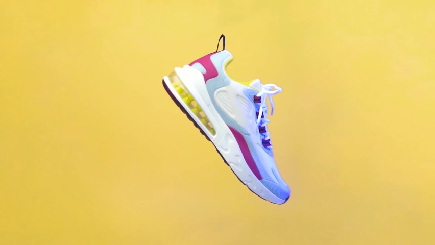 Spinning sports shoe concept, white shoes with a blend of colorful lines. Sport shoe rotation isolated on yellow background. Blue and red lighting from the side of the shoe. illustration of sport shoe Royalty-Free Stock Footage #1064545633