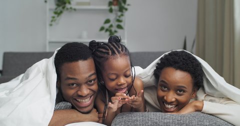 Happy family parents with cute child little daughter lie under blanket laugh hug look at camera. Mom father and child girl three people resting under blanket having fun spend time together at home