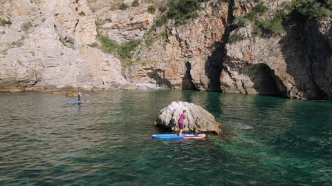Friends travel together and engage in active recreation. Man and woman row oars standing on SUP boards and swim along cliff. Beautiful sunny seascape with rock on background.