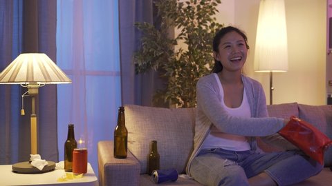 asian happy young woman gulping beverage from bottle and eating snacks while binge-watching episodes on tv alone at home.