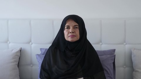 Portrait of sad and worried Asian senior Muslim woman, sitting on bed looking at camera, turning the face to the side ,and looking desperately. Handheld, close up shot, senior Asian Muslim concept