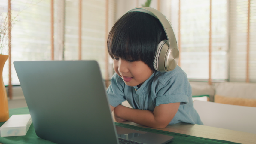 Little Cute Asian Boy in headphones having a video call talking using laptop sitting in the living room at home. Young child boy looking at laptop screen speaking online communication by video webcam Royalty-Free Stock Footage #1064564779